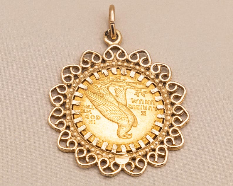 May include: A gold pendant with a coin featuring an eagle design, set in a decorative filigree frame. The pendant has a loop for a chain and is shown against a neutral backdrop.