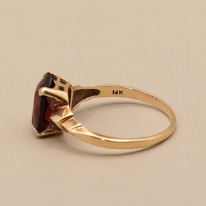 May include: A gold ring with a rectangular, dark red gemstone. The ring band is a simple, polished gold, with the marking "14K" visible. The gemstone is set in a gold bezel setting.