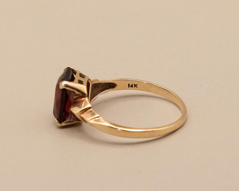 May include: A gold ring with a rectangular, dark red gemstone. The ring band is a simple, polished gold, with the marking "14K" visible. The gemstone is set in a gold bezel setting.