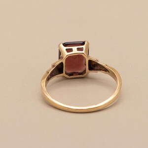 May include: A gold ring featuring a rectangular, reddish-brown gemstone. The ring's band is a simple, polished gold, and the gemstone is set in a gold bezel. The ring is viewed from a slightly elevated angle, showcasing its design and color.