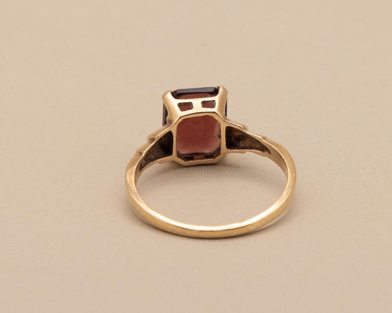 May include: A gold ring featuring a rectangular, reddish-brown gemstone. The ring's band is a simple, polished gold, and the gemstone is set in a gold bezel. The ring is viewed from a slightly elevated angle, showcasing its design and color.
