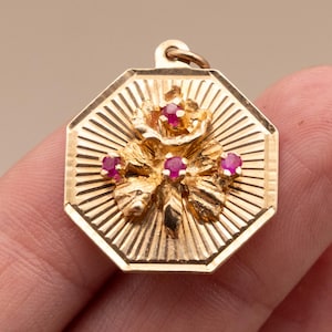 Könnte beinhalten: Goldfarbener Anhänger mit floralem Design und roten Edelsteinen. Der Anhänger hat eine achteckige Form mit einem strukturierten, sonnenstrahlenartigen Muster. Das florale Design zeigt eine Rose und Blätter, mit den Edelsteinen in der Mitte der Blumen.