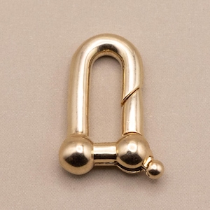 May include: A gold-colored, U-shaped shackle with a hinged opening. The shackle has a rounded top and two spherical ends. The metal hardware is designed for connecting items.