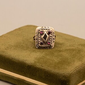 May include: A silver ring with a rectangular face, featuring a central diamond shape and small red gemstones. The ring is displayed on a green velvet surface, highlighting its detailed design. The ring's design includes decorative patterns around the edges.