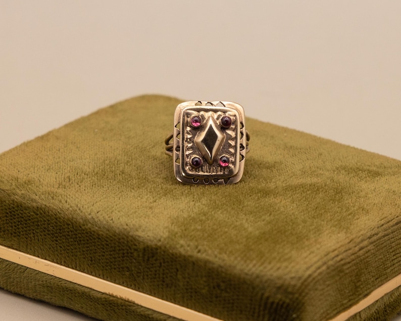 May include: A silver ring with a rectangular face, featuring a central diamond shape and small red gemstones. The ring is displayed on a green velvet surface, highlighting its detailed design. The ring's design includes decorative patterns around the edges.
