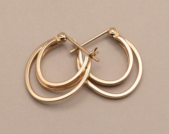 May include: A pair of gold hoop earrings featuring a double-layered design. Each earring consists of two concentric circular bands connected by a hinged post mechanism. The metal has a polished, reflective finish with a warm yellow gold tone.
