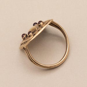 May include: A gold ring with an oval-shaped top, adorned with small, red gemstones. The ring's surface is textured, and the band is a simple, rounded design. The ring is a warm gold colour.