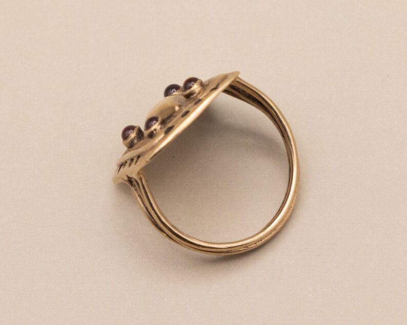 May include: A gold ring with an oval-shaped top, adorned with small, red gemstones. The ring's surface is textured, and the band is a simple, rounded design. The ring is a warm gold colour.