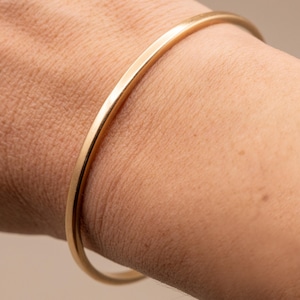 Op de afbeelding: Een gladde, goudkleurige armband met een afgerond profiel. De armband heeft een doorlopend, naadloos cirkelvormig ontwerp met een gepolijste metalen afwerking die het licht reflecteert langs het gebogen oppervlak.