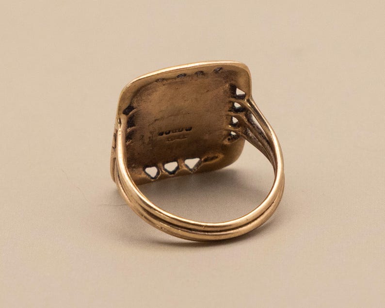 May include: A vintage gold ring with a rectangular face and a detailed band. The ring's face has a textured surface with small, decorative cutouts along the edges. The band features a double-layered design, adding to its classic appeal.