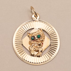 May include: A gold circular pendant featuring a textured cat figure in the center with two small green gemstone eyes. The cat is positioned on a diamond-shaped plate within a round frame that has a sunburst-style etched pattern along the edge.