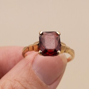 May include: A gold ring featuring a large, rectangular, deep red gemstone. The ring's band has a textured design, and the gemstone is held in place by four gold prongs. The ring is held by a person's fingers.