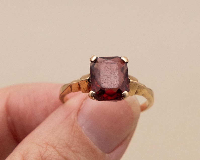 May include: A gold ring featuring a large, rectangular, deep red gemstone. The ring's band has a textured design, and the gemstone is held in place by four gold prongs. The ring is held by a person's fingers.