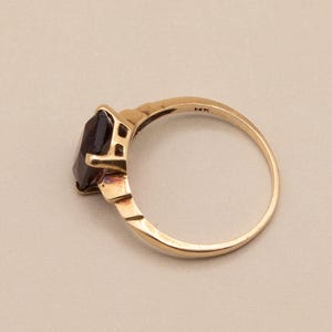 May include: A gold ring with a dark gemstone. The ring has a simple band and a rectangular setting that holds the dark gemstone. The ring is marked "14K". The ring is shown on a neutral background.