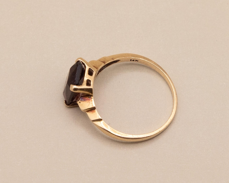 May include: A gold ring with a dark gemstone. The ring has a simple band and a rectangular setting that holds the dark gemstone. The ring is marked "14K". The ring is shown on a neutral background.