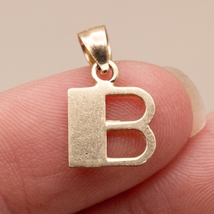 May include: A gold-toned metal pendant shaped as the capital letter B. The pendant features a flat, block-style font with a smooth surface and a small, integrated circular bail at the top for attachment to a chain or necklace cord.