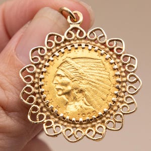 May include: A gold pendant featuring a coin with an image of a Native American in a headdress. The coin is set in a decorative, openwork frame. The pendant is held by a hand.