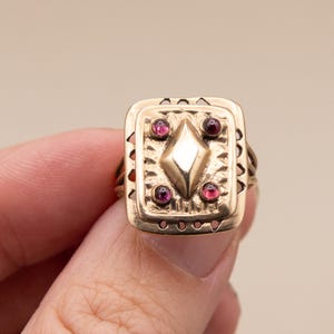 May include: A gold ring with a rectangular face. The face features a central diamond shape and four surrounding red gemstones. The ring has an ornate, vintage design with decorative edges and detailing.