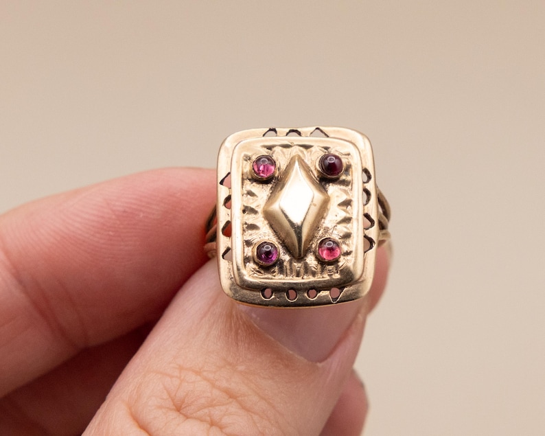May include: A gold ring with a rectangular face. The face features a central diamond shape and four surrounding red gemstones. The ring has an ornate, vintage design with decorative edges and detailing.