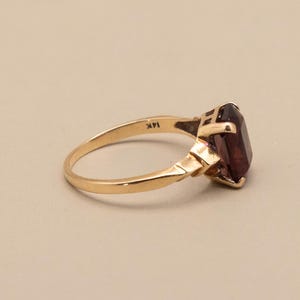 May include: A gold ring with a large, rectangular, dark red gemstone. The ring band is a polished gold color, and the setting has a geometric design. The ring is marked "14K".