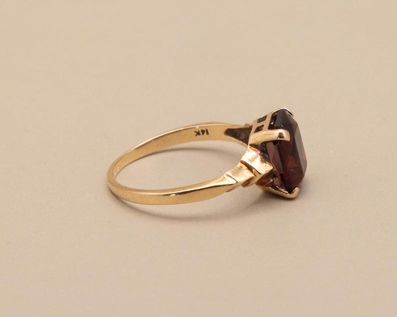 May include: A gold ring with a large, rectangular, dark red gemstone. The ring band is a polished gold color, and the setting has a geometric design. The ring is marked "14K".