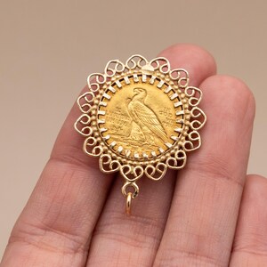 May include: A gold pendant featuring a coin with an eagle design, set within a decorative, openwork frame. The pendant has a small loop at the bottom for attachment. The coin is a warm gold colour.