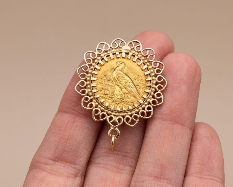 May include: A gold pendant featuring a coin with an eagle design, set within a decorative, openwork frame. The pendant has a small loop at the bottom for attachment. The coin is a warm gold colour.