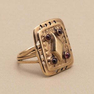 May include: A gold ring with a rectangular face. The face has a diamond shape and three small, round, red gemstones. The ring has a detailed design with small cut-out patterns and a triple-banded shank.