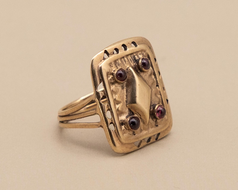 May include: A gold ring with a rectangular face. The face has a diamond shape and three small, round, red gemstones. The ring has a detailed design with small cut-out patterns and a triple-banded shank.