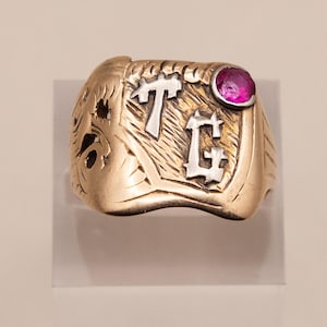 Può includere: Un anello color oro con una superficie testurizzata e le lettere T e G color argento ben visibili. L'anello presenta una piccola pietra preziosa rotonda di colore rosa acceso incastonata nell'angolo in alto a destra, oltre a dettagli incisi.
