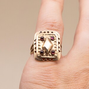 May include: A gold ring with a square face, featuring a central diamond shape and four red gemstones. The ring has a textured design and is worn on a finger.