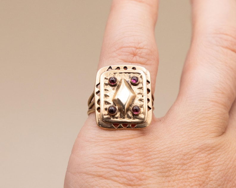 May include: A gold ring with a square face, featuring a central diamond shape and four red gemstones. The ring has a textured design and is worn on a finger.