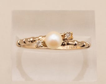 May include: A gold ring featuring a central cream-colored pearl flanked by two small, clear round-cut stones. The band has a textured, ribbed design that tapers toward the center, creating a bypass-style setting with a polished metallic finish.