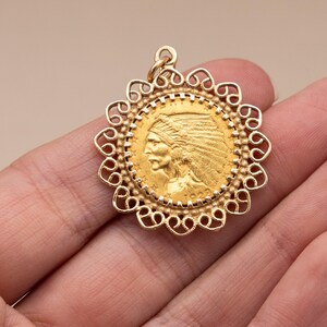 May include: A gold pendant with a detailed profile of a Native American chief, set within an ornate, openwork frame. The pendant has a loop for a chain and is likely a piece of jewellery. The coin-like design suggests a vintage or antique style.