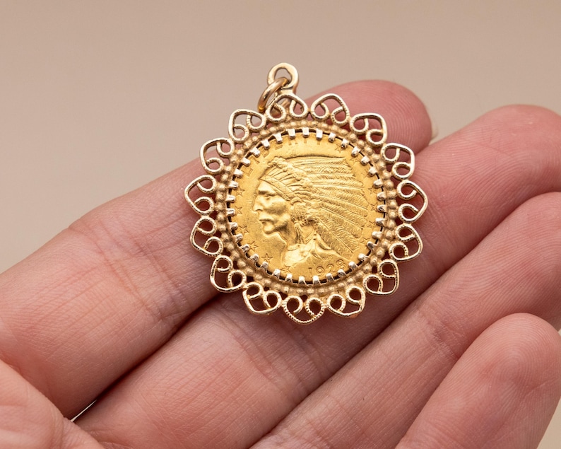 May include: A gold pendant with a detailed profile of a Native American chief, set within an ornate, openwork frame. The pendant has a loop for a chain and is likely a piece of jewellery. The coin-like design suggests a vintage or antique style.
