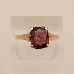 May include: A gold ring featuring a large, rectangular, dark red gemstone. The ring's band has a textured design near the gemstone setting. The gemstone is the focal point, showcasing its deep color and cut.