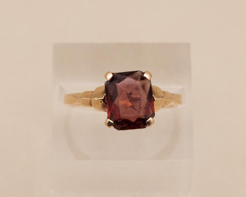 May include: A gold ring featuring a large, rectangular, dark red gemstone. The ring's band has a textured design near the gemstone setting. The gemstone is the focal point, showcasing its deep color and cut.