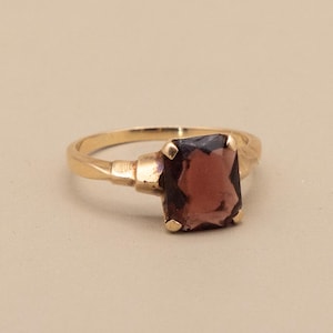 May include: A gold ring featuring a large, rectangular, dark red gemstone. The ring has a simple band with decorative elements near the gemstone setting. The gemstone is the focal point, catching the light and adding a touch of elegance.