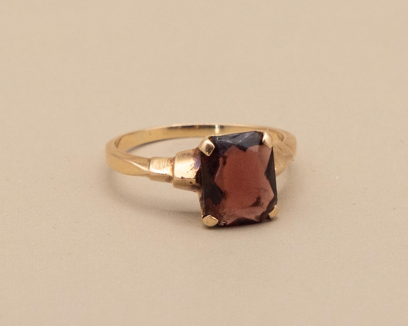 May include: A gold ring featuring a large, rectangular, dark red gemstone. The ring has a simple band with decorative elements near the gemstone setting. The gemstone is the focal point, catching the light and adding a touch of elegance.