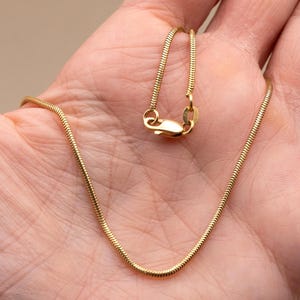 Puede incluir: Un collar de cadena tipo serpiente en tono dorado con un diseño de eslabones redondeados, lisos y flexibles. El collar incluye un mecanismo de cierre de mosquetón seguro en el extremo, ofreciendo un aspecto pulido y uniforme.