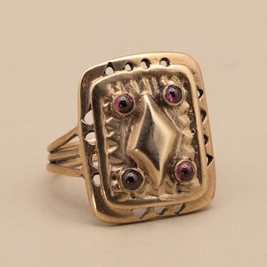 May include: A gold-coloured ring with a rectangular face. The face features a diamond shape in the centre, surrounded by four small, round, red gemstones. The ring has a detailed, textured design and a split band.