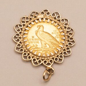 May include: A gold pendant with a gold coin at its centre, featuring an eagle design. The coin is set in an ornate, openwork frame. The coin bears the inscriptions "E PLURIBUS UNUM" and "IN GOD WE TRUST". A small loop is attached.