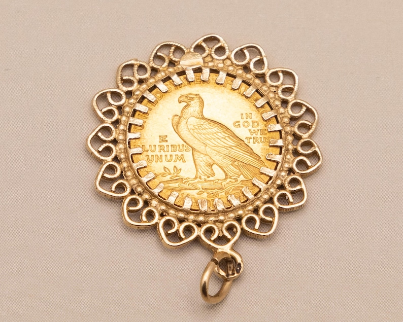 May include: A gold pendant with a gold coin at its centre, featuring an eagle design. The coin is set in an ornate, openwork frame. The coin bears the inscriptions "E PLURIBUS UNUM" and "IN GOD WE TRUST". A small loop is attached.