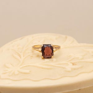May include: A gold ring with a rectangular, dark red gemstone. The ring is set on a cream-colored, heart-shaped box with floral carvings. The ring's band is thin and simple, with a classic design.