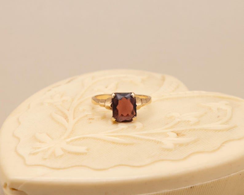 May include: A gold ring with a rectangular, dark red gemstone. The ring is set on a cream-colored, heart-shaped box with floral carvings. The ring's band is thin and simple, with a classic design.