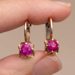 May include: A pair of gold-toned earrings with a lever back closure. Each earring features a vibrant, round, magenta-colored gemstone set in a four-petal floral design. The earrings are held between fingers.