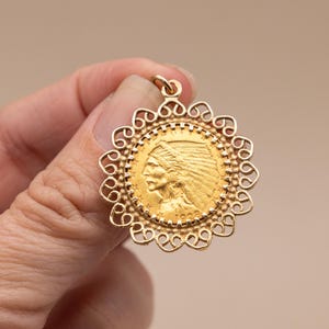 May include: A gold pendant with a detailed profile of a Native American in a headdress, surrounded by a decorative filigree border. The pendant has a loop for a chain and is held in a hand.
