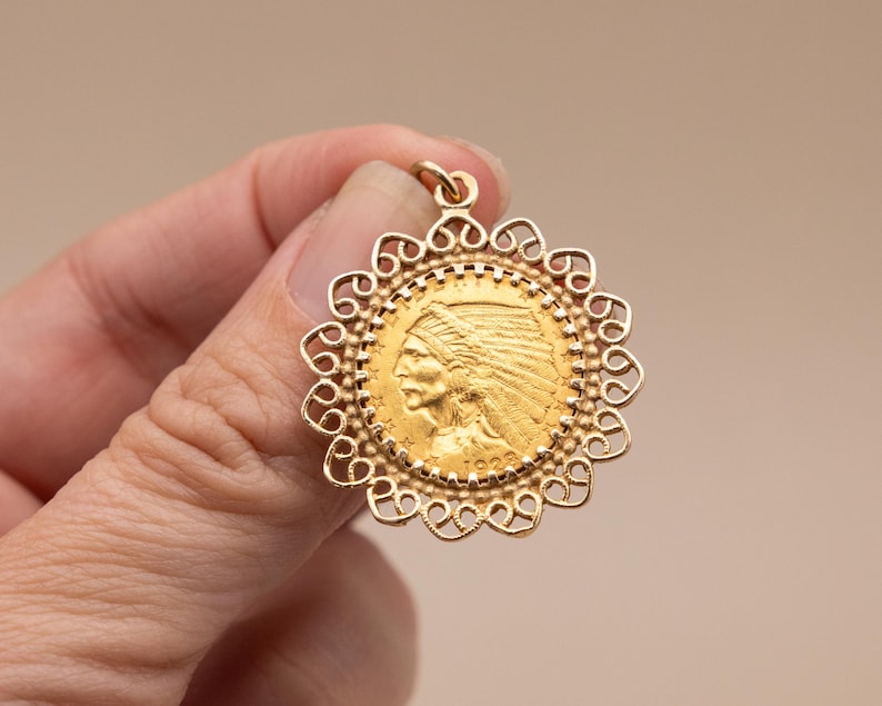 May include: A gold pendant with a detailed profile of a Native American in a headdress, surrounded by a decorative filigree border. The pendant has a loop for a chain and is held in a hand.