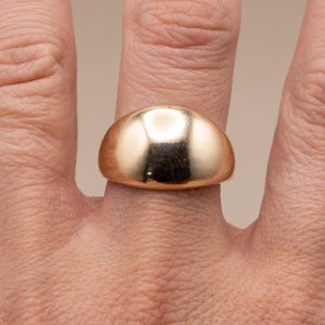 Può includere: Un anello color oro con un design arrotondato e bombato. L'anello è indossato su un dito e la superficie lucida riflette la luce. Il design semplice ma elegante dell'anello lo rende un accessorio versatile.