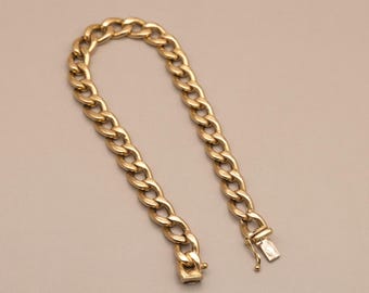 May include: A gold-toned metal chain bracelet featuring interlocking oval links. The bracelet is arranged in a curved shape and includes a secure box clasp mechanism at one end, showing a polished finish throughout the entire piece.